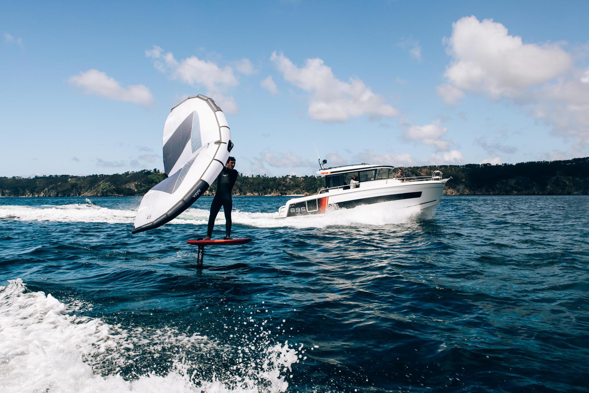 a person on a surfboard with a sail on the water with a boat Merry fisher 895 S2 in the background